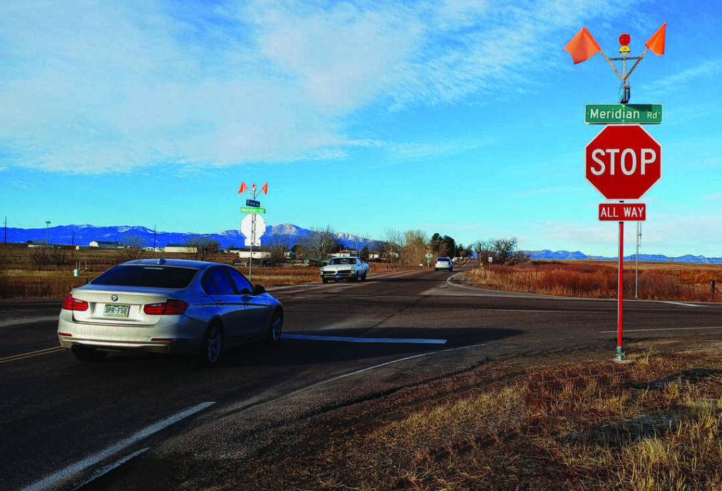 A car waits at a stop sign at the intersection of Meridian Road, with mountains visible in the background under a partly cloudy sky.