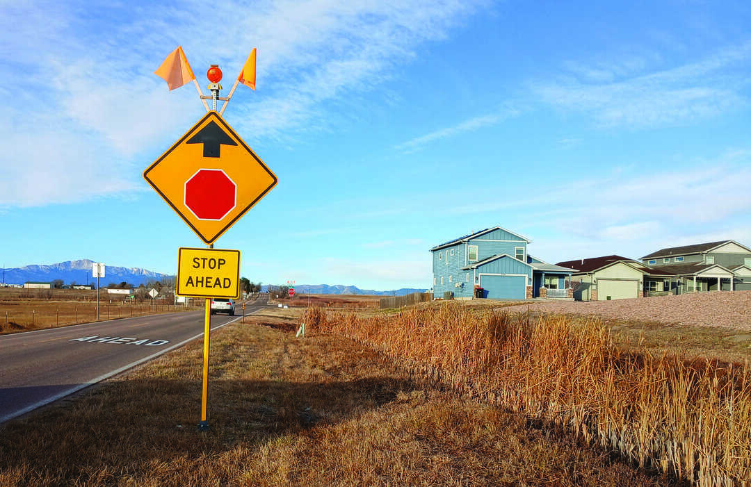 Yellow "STOP AHEAD" sign and stop warning sign with orange flags beside a rural road, houses and dry grass field visible on the right.