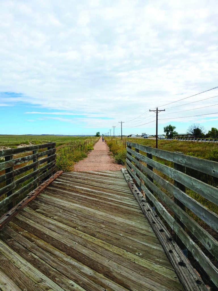 A wooden bridge leads to a straight gravel path that stretches into the distance through open fields under a partly cloudy sky.