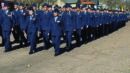 A large group of uniformed military personnel marches in formation on a paved outdoor area under a clear sky.