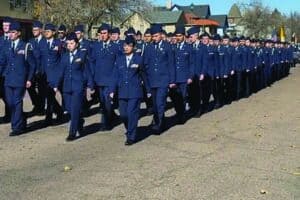 A large group of uniformed military personnel marches in formation on a paved outdoor area under a clear sky.