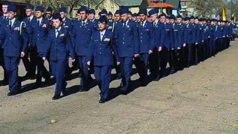 A large group of uniformed military personnel marches in formation on a paved outdoor area under a clear sky.