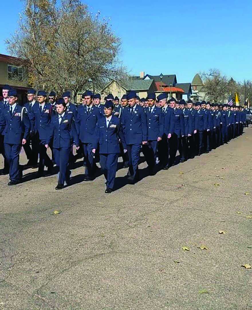 A large group of uniformed military personnel marches in formation on a paved outdoor area under a clear sky.