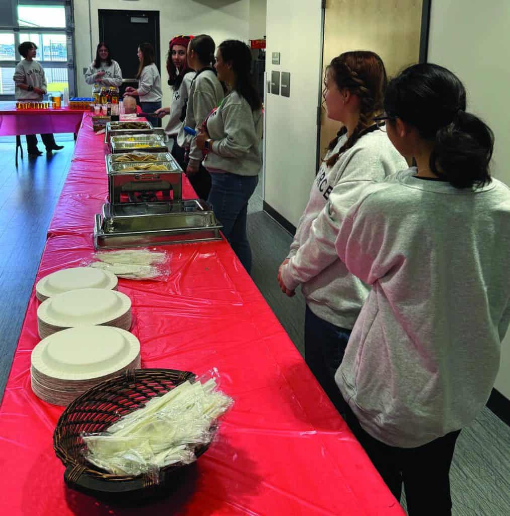 A group of people in gray shirts stand by a long table with a red tablecloth, set with plates, utensils, and trays of food in a room.