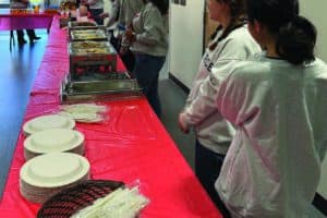 A group of people in gray shirts stand by a long table with a red tablecloth, set with plates, utensils, and trays of food in a room.