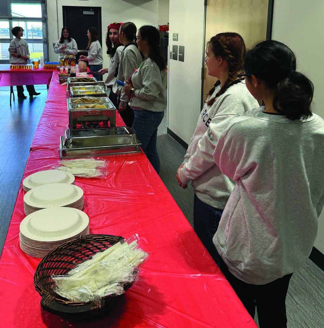 A group of people in gray shirts stand by a long table with a red tablecloth, set with plates, utensils, and trays of food in a room.
