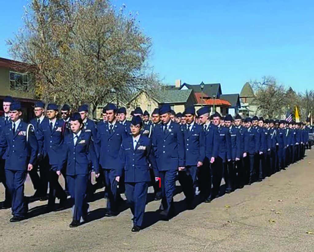 A large group of people in blue military uniforms march in formation down a street lined with trees and houses.