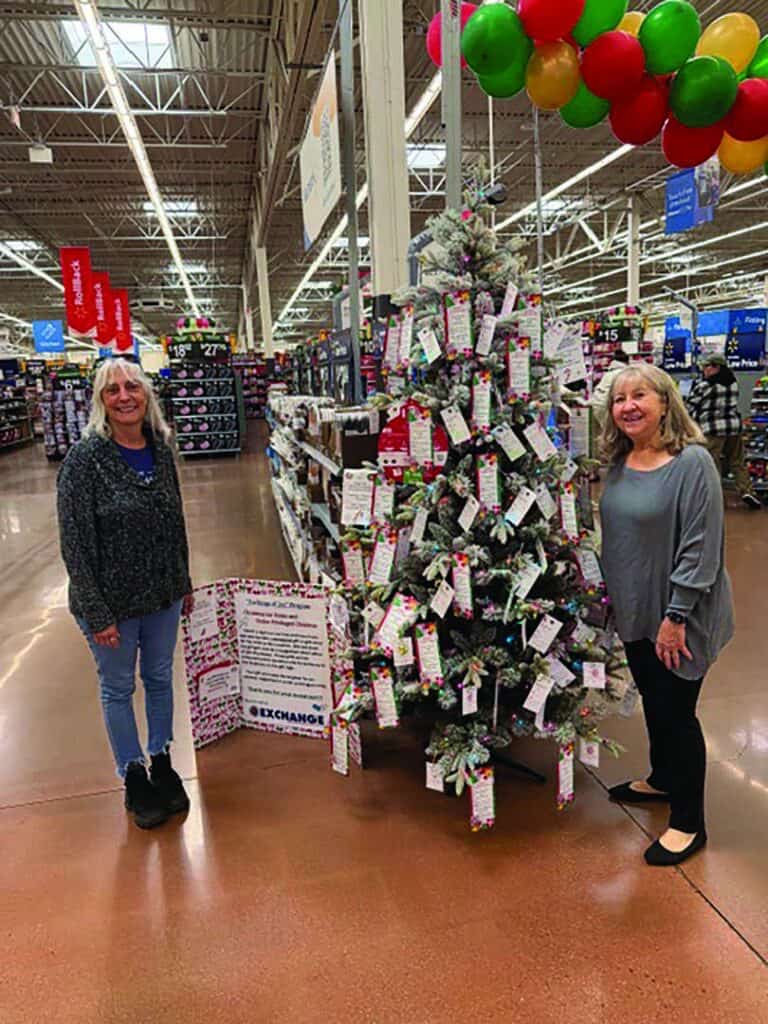 Two women stand beside a decorated Christmas tree with tags in a store, under colorful balloons, with store aisles and a sign visible in the background.