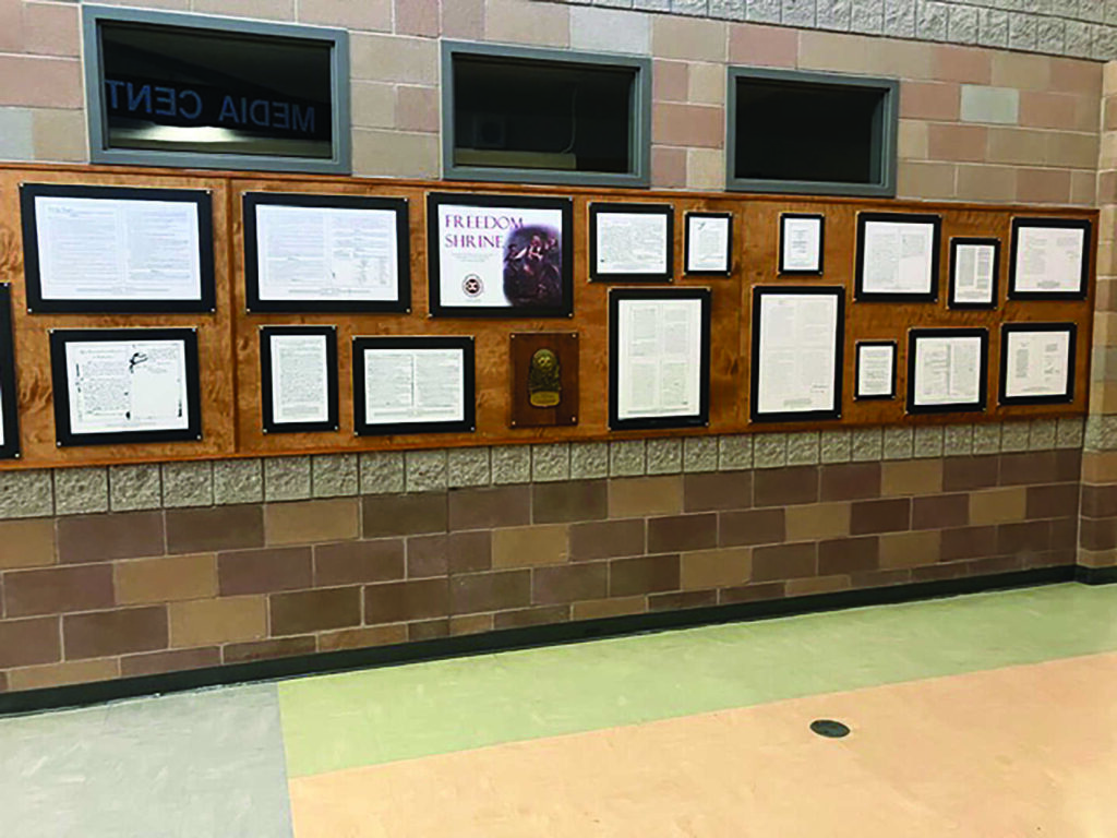 A bulletin board displays framed documents and a poster titled "Freedom Shrine" on a brick wall in a hallway with tiled flooring.