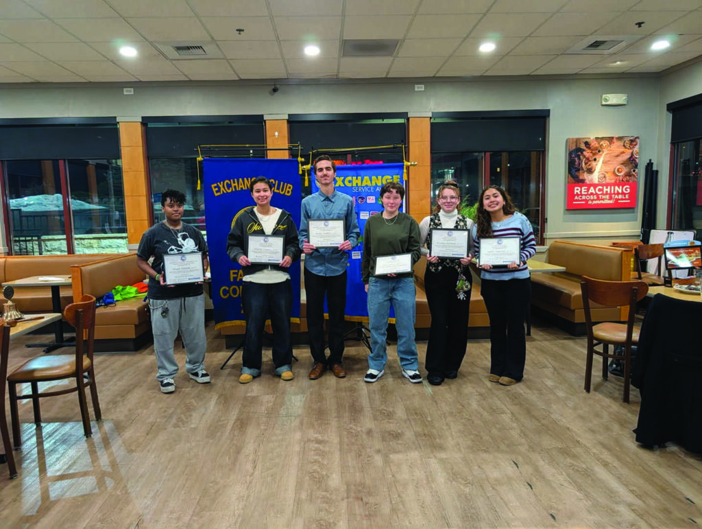 Six people stand indoors holding certificates in front of Exchange Club banners, posing for a group photo in a restaurant setting.