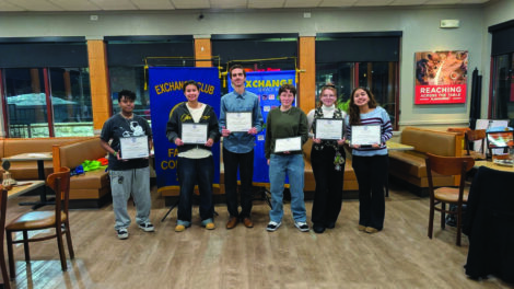 Six people stand indoors holding certificates in front of Exchange Club banners, posing for a group photo in a restaurant setting.
