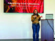 A woman stands at a podium giving a presentation titled "Advancing Food is Medicine: Healing People & the Land" with a FreshRx Oklahoma logo displayed on a screen behind her.