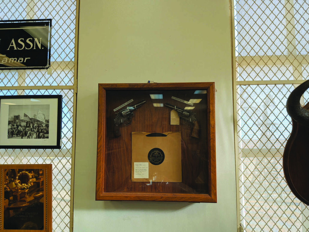 A wooden display case with two pistols and a sealed envelope is mounted on a wall between two barred windows. Framed photos and plaques surround the case.
