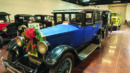 A row of vintage cars is displayed indoors; the front car is blue with a festive wreath and bow attached to its grille.