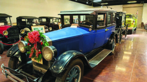 A row of vintage cars is displayed indoors; the front car is blue with a festive wreath and bow attached to its grille.