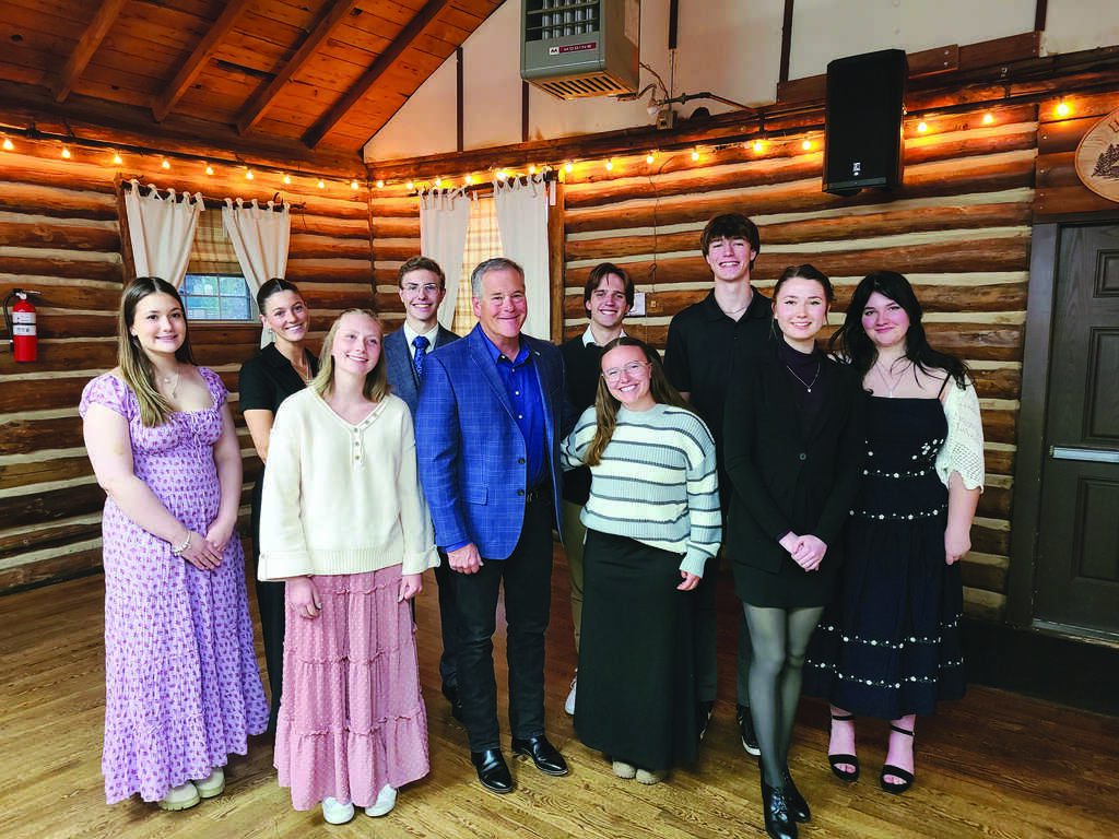 A group of ten people, dressed in semi-formal attire, stands together smiling in a wooden log cabin room with string lights overhead.