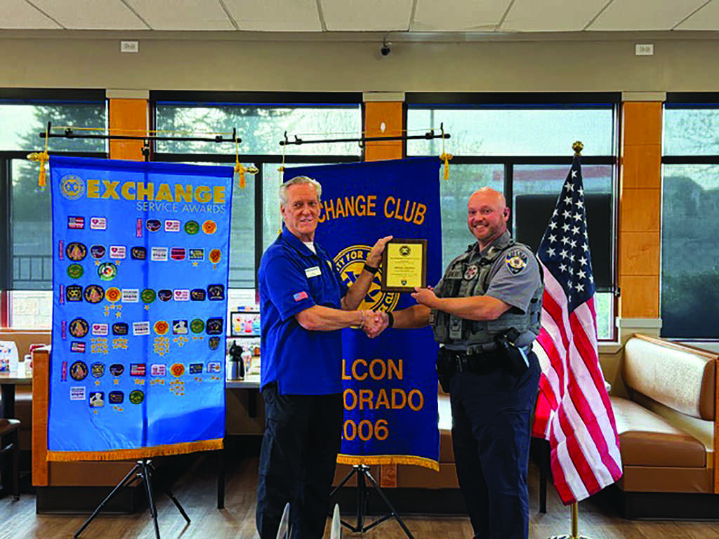 Two men stand indoors shaking hands as one hands a plaque to the other. Behind them are banners for the Exchange Club and a U.S. flag.