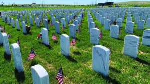 Rows of white gravestones with small American flags placed beside them in a cemetery on a sunny day.