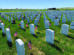 Rows of white gravestones with small American flags placed beside them in a cemetery on a sunny day.
