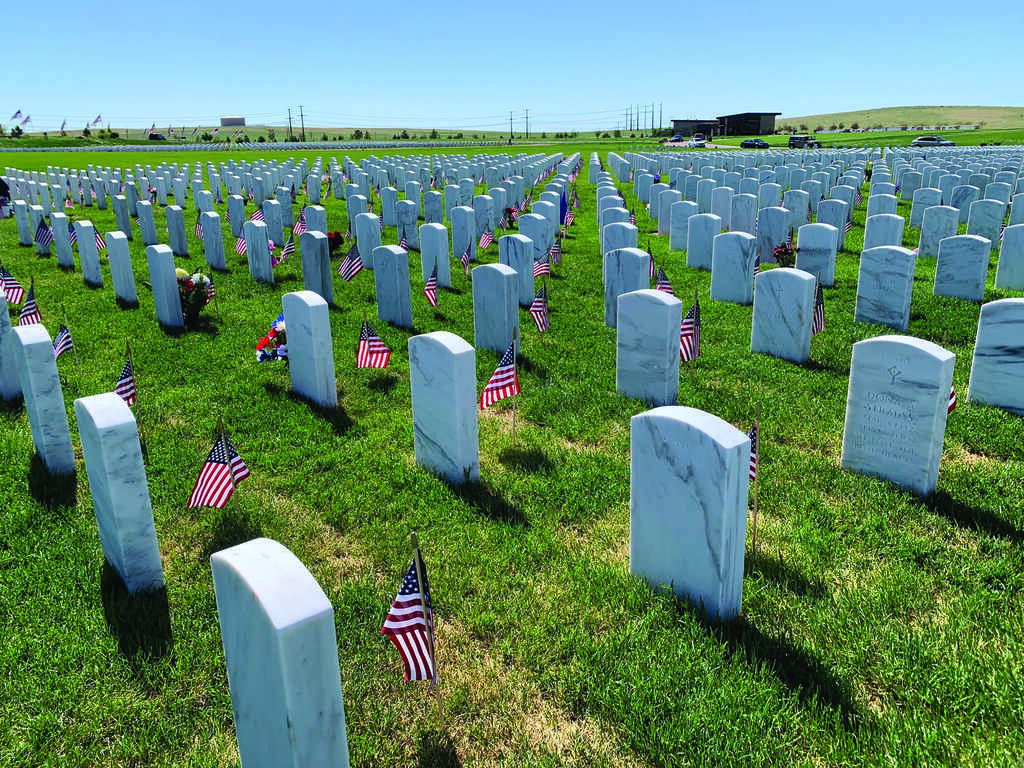 Rows of white gravestones with small American flags placed beside them in a cemetery on a sunny day.