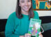 A woman in a green sweater sits on a couch, smiling and holding a children’s book titled "Into the Field" with an illustrated cover.