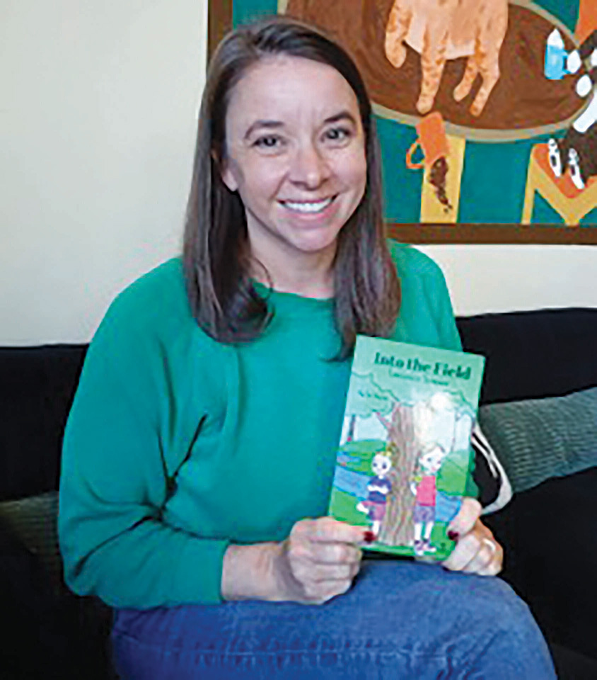 A woman in a green sweater sits on a couch, smiling and holding a children’s book titled "Into the Field" with an illustrated cover.