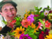 A person wearing a green cap smiles while holding a large, colorful bouquet of assorted flowers in front of a plain background.