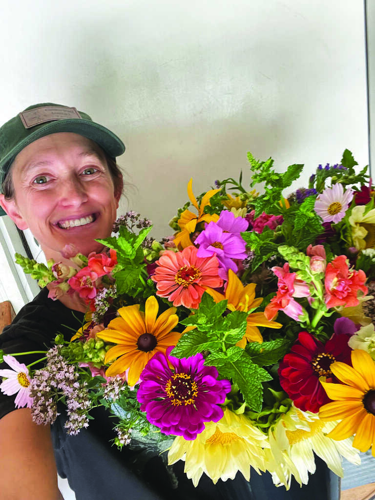 A person wearing a green cap smiles while holding a large, colorful bouquet of assorted flowers in front of a plain background.