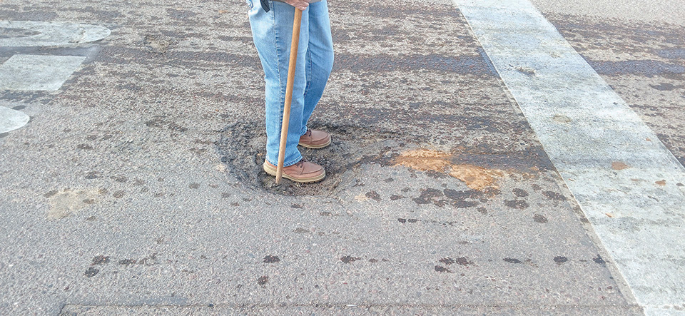 Person standing in a pothole on an asphalt road near a crosswalk, holding a stick and wearing jeans and brown shoes.