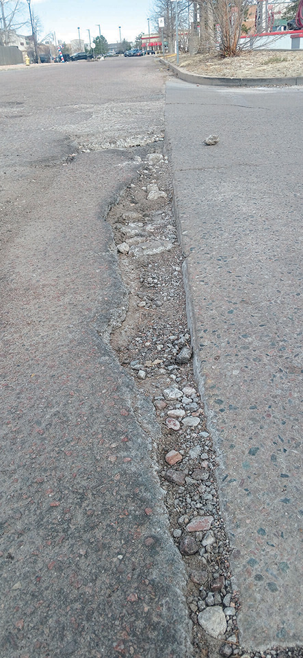 A close-up view of a road with a large pothole and crumbling asphalt along the curb, with some debris scattered nearby.