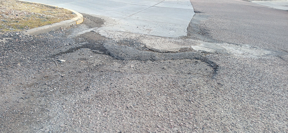 Damaged road surface with cracks and uneven patches near a concrete curb, showing wear and signs of disrepair.