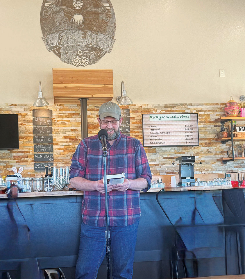 A man in a plaid shirt and cap stands at a microphone reading from a notepad in front of a bar with a menu board and decorative sign behind him.