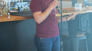 A woman stands beside a bar counter, holding a microphone and reading from a sheet of paper in a casual indoor setting.