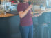 A woman stands beside a bar counter, holding a microphone and reading from a sheet of paper in a casual indoor setting.
