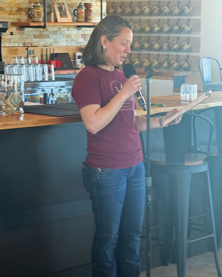A woman stands beside a bar counter, holding a microphone and reading from a sheet of paper in a casual indoor setting.