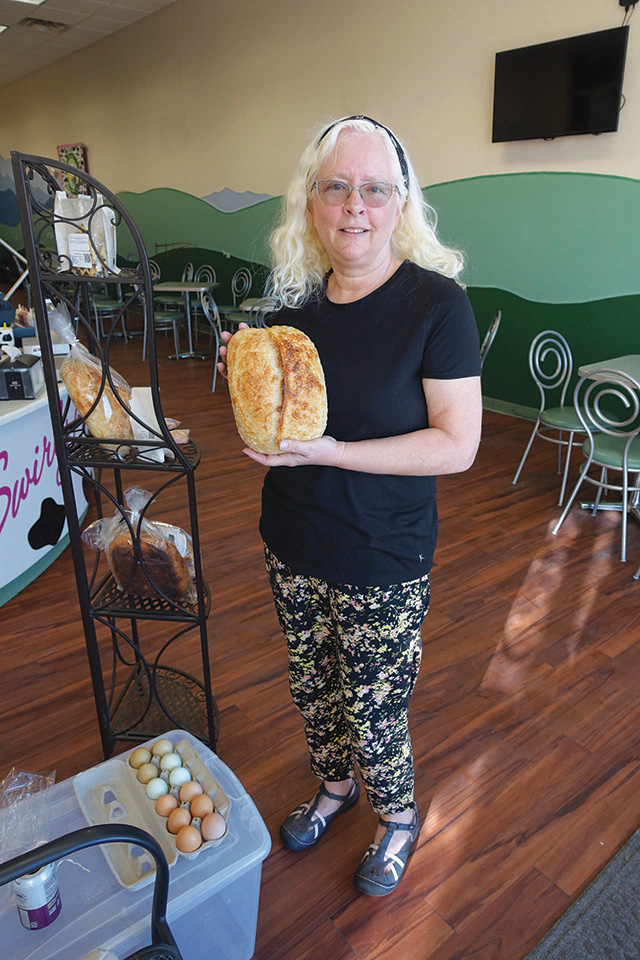 A woman with light hair and glasses holds a loaf of bread in a cafe. A shelf with bread and a container of eggs are nearby.