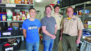 Four men stand and smile in a food pantry, with shelves of packaged food and supplies visible behind them. Two are in scout uniforms, one wears a blue shirt, and one wears a gray shirt.