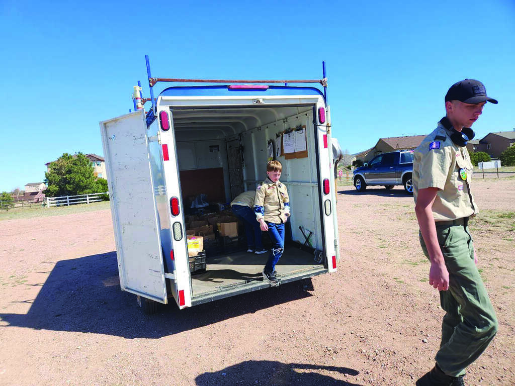 Two boys in scouting uniforms unload supplies from a white trailer parked in a dirt lot on a sunny day.