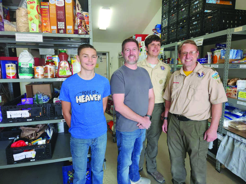 Four men stand and smile in a food pantry, with shelves of packaged food and supplies visible behind them. Two are in scout uniforms, one wears a blue shirt, and one wears a gray shirt.