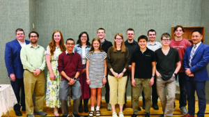 A group of thirteen people, including adults and teenagers, stand indoors on a wooden floor, posing for a group photo against a gray wall.