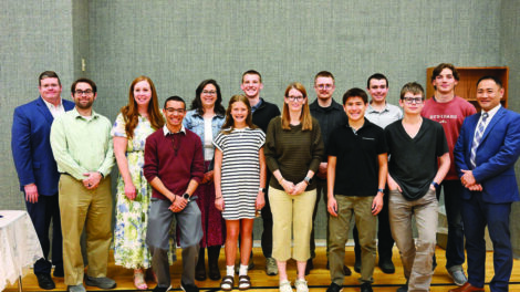 A group of thirteen people, including adults and teenagers, stand indoors on a wooden floor, posing for a group photo against a gray wall.