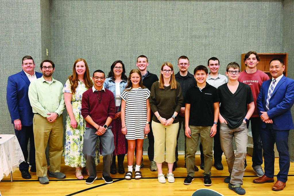 A group of thirteen people, including adults and teenagers, stand indoors on a wooden floor, posing for a group photo against a gray wall.