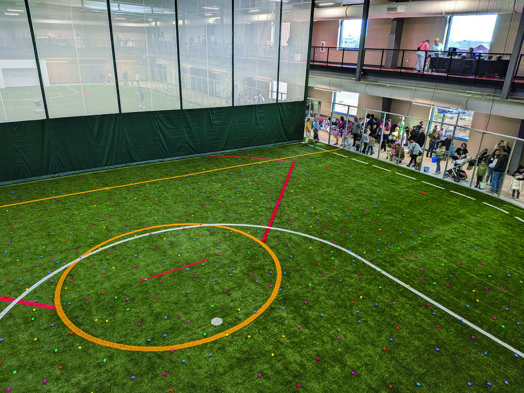 Indoor turf field with small colored objects scattered across it, a crowd of people lined up along the right side, and an upper level visible in the background.