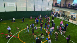 Children and adults gather on artificial turf inside a large indoor sports facility, participating in an egg hunt event with colorful eggs scattered on the ground.