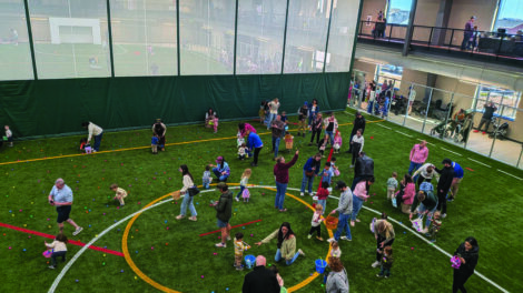 Children and adults gather on artificial turf inside a large indoor sports facility, participating in an egg hunt event with colorful eggs scattered on the ground.