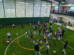 Children and adults gather on artificial turf inside a large indoor sports facility, participating in an egg hunt event with colorful eggs scattered on the ground.