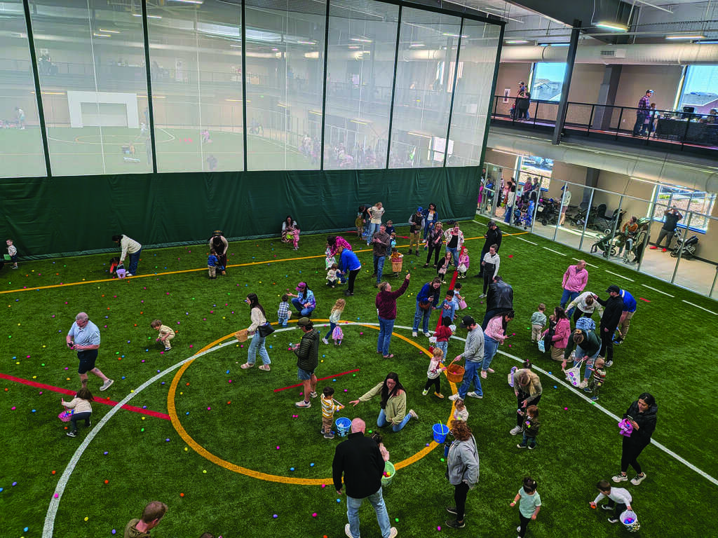 Children and adults gather on artificial turf inside a large indoor sports facility, participating in an egg hunt event with colorful eggs scattered on the ground.