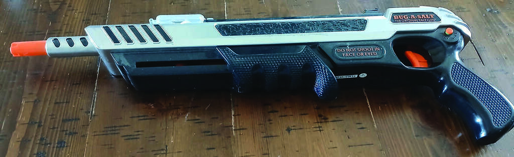 A Bug-A-Salt plastic gun with an orange tip rests on a wooden surface. The device is designed for shooting salt to kill insects.