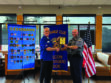 Two men shake hands as one presents an award plaque to the other, standing in front of banners and an American flag in a restaurant setting.
