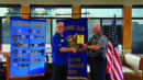 Two men shake hands as one presents an award plaque to the other, standing in front of banners and an American flag in a restaurant setting.